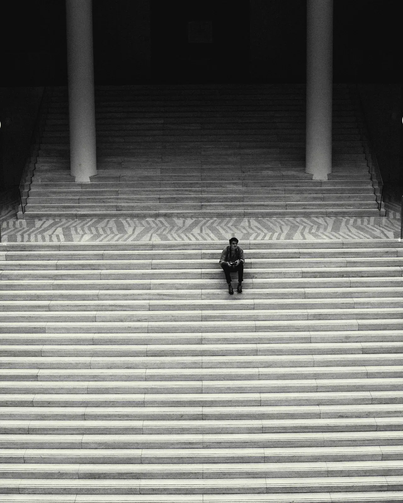 A single person sitting on a wide flight of concrete steps between two columns.